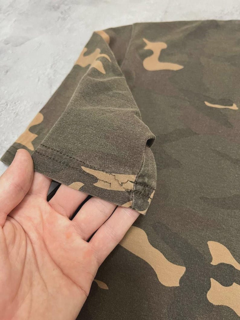 Puede incluir: Primer plano de una camiseta con estampado de camuflaje. La camiseta presenta una mezcla de colores verde oliva, marr&oacute;n y beige. La tela parece ser un material suave, posiblemente de algod&oacute;n. La manga es visible, con el borde de la manga enfocado.