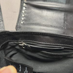 Puede incluir: Bolso de mano de cuero negro con tachuelas plateadas en la solapa. El interior tiene un bolsillo con cremallera y una peque&ntilde;a etiqueta. El bolso est&aacute; abierto, revelando el forro y un cierre de cremallera.