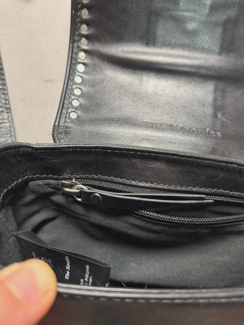 Puede incluir: Bolso de mano de cuero negro con tachuelas plateadas en la solapa. El interior tiene un bolsillo con cremallera y una peque&ntilde;a etiqueta. El bolso est&aacute; abierto, revelando el forro y un cierre de cremallera.