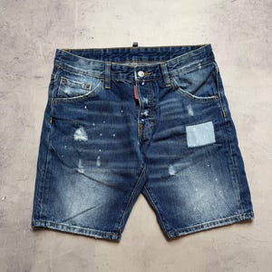 Może przedstawiać: Niebieskie szorty dżinsowe z zapięciem na guziki, przetarciami i jasnoniebieską naszywką. Szorty mają klasyczny pięciokieszeniowy design i są lekko wyblakłe z białymi plamami farby.