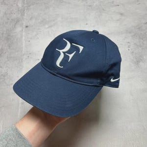 Può includere: Cappellino da baseball blu navy con logo "RF" bianco ricamato sul davanti. Il cappellino ha una visiera curva e un piccolo logo Nike bianco sul lato. Realizzato in tessuto resistente.