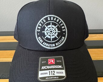 May include: A black trucker hat with a white logo that says "Capts Quarters Bene Armatum Militiam" and a white compass design. The hat has a black mesh back and a black adjustable strap with a white label that says "Richardson® Original 112 Trucker Adjustable".