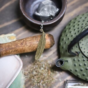 May include: A green cast iron teapot with a black handle and a gold feather tea infuser. The teapot is sitting on a wooden surface.