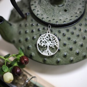 May include: A silver tree of life charm pendant hangs from a green cast iron teapot. The teapot has a textured surface and a round lid.