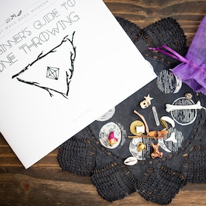 May include: A beginner's guide to bone throwing, a booklet with a black and white illustration of a triangle with a diamond inside. A purple pouch with a drawstring sits on a black lace doily with a moon and star pattern. The pouch contains a variety of small bones, crystals, and other objects.