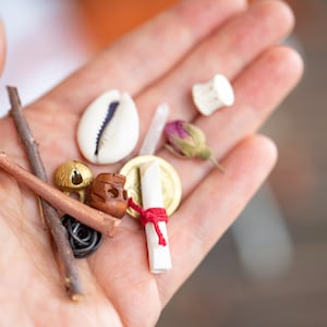 May include: A hand holding a collection of small objects, including a seashell, a small wooden figure, a rolled up piece of paper tied with red string, a small gold coin, a twig, and a dried rose bud.