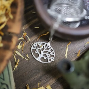 Puede incluir: Un colgante de plata en forma de árbol de la vida con detalles intrincados. El colgante está sobre una superficie de madera con pétalos de flores secas.