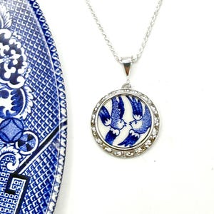 Peut inclure: Un collier pendentif circulaire composé d'un éclat de céramique bleu et blanc orné d'un motif d'oiseaux, serti dans une monture en métal argenté au bord texturé, suspendu à une fine chaîne argentée, présenté près d'une assiette à motifs bleus.