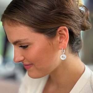 Peut inclure: Une boucle d'oreille cr&eacute;ole en argent avec un pendentif circulaire incrust&eacute; de nacre blanche iris&eacute;e et un petit embl&egrave;me d'abeille dor&eacute; au centre. Le bijou pr&eacute;sente un bord perl&eacute; textur&eacute; autour du m&eacute;daillon circulaire.