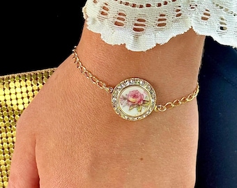 Peut inclure: Un bracelet à chaîne dorée doté d'un pendentif circulaire central orné d'un motif floral représentant une rose rose sur fond blanc, entouré d'une bordure de petits cristaux clairs. La fine chaîne métallique est reliée au sertissage circulaire.
