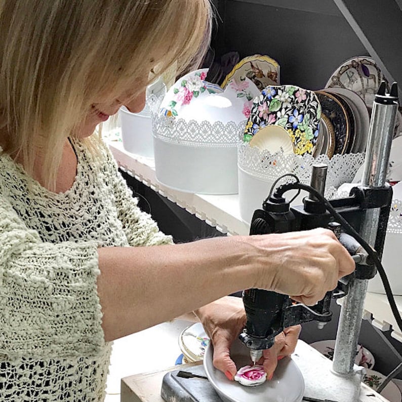 May include: A person using a drill press to work on a small, white ceramic piece with a pink rose design. White decorative boxes with floral patterns and lace trim are visible on a shelf in the background. The scene suggests a craft or artisan workshop.
