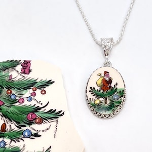 May include: Silver-toned necklace with an oval pendant featuring a vintage illustration of Santa Claus with a sack of toys. The pendant has a decorative silver border and a white background. A fragment of a Christmas tree is visible.