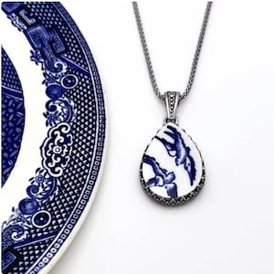 May include: A teardrop-shaped pendant necklace with a white and blue bird design. The pendant is framed with a decorative silver-tone metal. A matching blue and white plate is in the background.