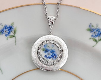 May include: A silver-toned pendant necklace with a round locket. The locket features a floral design with blue flowers and a crystal border. The chain is delicate and the locket has an engraved floral pattern around the edge.