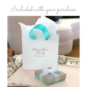 May include: A white gift bag with a teal ribbon handle and tissue paper. The bag has the text "Dinner Wear Jewelry" and "Wear A Piece of the Past". A small gift box with a white ribbon is also included.