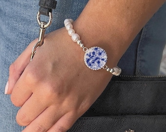 Könnte beinhalten: Ein Armband mit weißen Perlen und einem zentralen runden Anhänger mit blauem Blumenmuster. Der Anhänger ist in silberfarbenem Metall gefasst. Das Armband wird am Handgelenk getragen, mit einer schwarzen Handtasche und Jeans im Hintergrund.