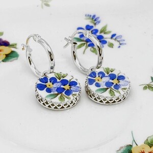 Peut inclure: Une paire de boucles d'oreilles cr&eacute;oles en argent avec des pendentifs circulaires blancs orn&eacute;s d'un motif floral bleu et jaune. Chaque pendentif est serti dans une monture argent&eacute;e festonn&eacute;e, suspendu &agrave; un anneau en argent poli.