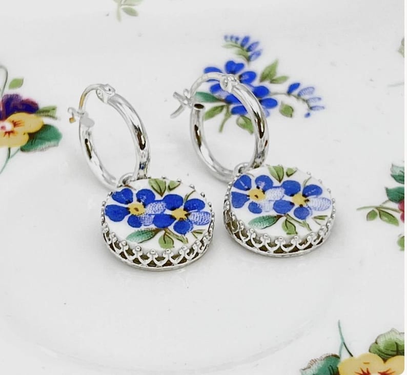 Peut inclure: Une paire de boucles d'oreilles cr&eacute;oles en argent avec des pendentifs circulaires blancs orn&eacute;s d'un motif floral bleu et jaune. Chaque pendentif est serti dans une monture argent&eacute;e festonn&eacute;e, suspendu &agrave; un anneau en argent poli.