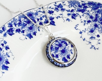 Könnte beinhalten: Eine silberne Anhängerkette mit einem kreisförmigen Mittelstück, das ein blau-weißes Blumenmuster aufweist und von einem Ring aus kleinen blauen Kristallen umgeben ist. Der Anhänger hängt an einer dünnen Silberkette, passend zum Blumenmuster.