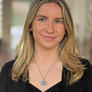 May include: A silver necklace with a circular pendant displaying a blue and white design. The pendant is encircled by small, clear stones. The necklace is worn by a person with blonde hair and a black blazer.