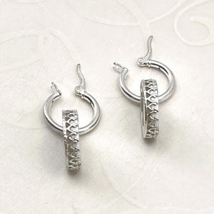 Peut inclure: Une paire de cr&eacute;oles en argent, chacune compos&eacute;e d'un petit anneau poli avec fermoir &agrave; charni&egrave;re et d'une breloque circulaire plus &eacute;paisse orn&eacute;e d'un motif r&eacute;p&eacute;titif de c&oelig;urs ou de fleurs de lys d&eacute;coup&eacute;s.