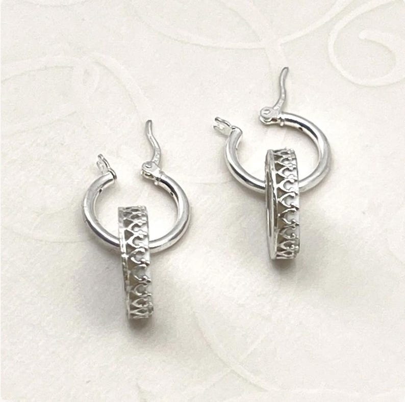 Peut inclure: Une paire de cr&eacute;oles en argent, chacune compos&eacute;e d'un petit anneau poli avec fermoir &agrave; charni&egrave;re et d'une breloque circulaire plus &eacute;paisse orn&eacute;e d'un motif r&eacute;p&eacute;titif de c&oelig;urs ou de fleurs de lys d&eacute;coup&eacute;s.