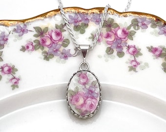 May include: Silver-toned necklace with an oval pendant featuring a floral design in shades of pink and purple. The pendant is set against a white background and framed with a decorative border. The necklace is displayed on a floral patterned plate.