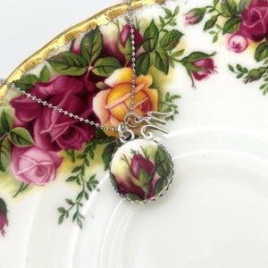May include: A silver necklace with a round pendant featuring a floral design of red and yellow roses. The pendant is set against a white background and is on a white plate with a floral pattern.