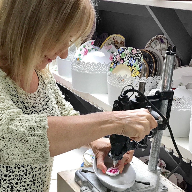 K&ouml;nnte beinhalten: Eine Person verwendet eine St&auml;nderbohrmaschine, um an einem kleinen, dekorativen Keramikst&uuml;ck mit einem rosa Rosenmuster zu arbeiten. Im Hintergrund sind Regale mit wei&szlig;en Beh&auml;ltern und dekorativen Tellern zu sehen.