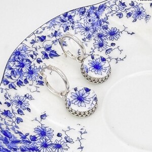 Peut inclure: Une paire de boucles d'oreilles cr&eacute;oles en argent avec des pendentifs circulaires. Chaque pendentif pr&eacute;sente un fond blanc orn&eacute; d'un motif floral bleu, entour&eacute; d'une monture en argent festonn&eacute;e assortie aux cr&eacute;oles.