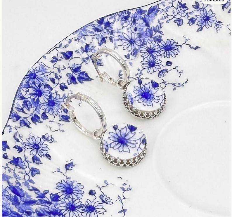 Peut inclure: Une paire de boucles d'oreilles cr&eacute;oles en argent avec des pendentifs circulaires. Chaque pendentif pr&eacute;sente un fond blanc orn&eacute; d'un motif floral bleu, entour&eacute; d'une monture en argent festonn&eacute;e assortie aux cr&eacute;oles.