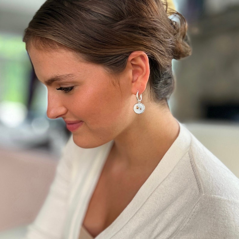 Peut inclure: Une boucle d'oreille cr&eacute;ole en argent avec un pendentif circulaire incrust&eacute; de nacre blanche iris&eacute;e et un petit embl&egrave;me d'abeille dor&eacute; au centre. Le bijou pr&eacute;sente un bord perl&eacute; textur&eacute; autour du m&eacute;daillon circulaire.