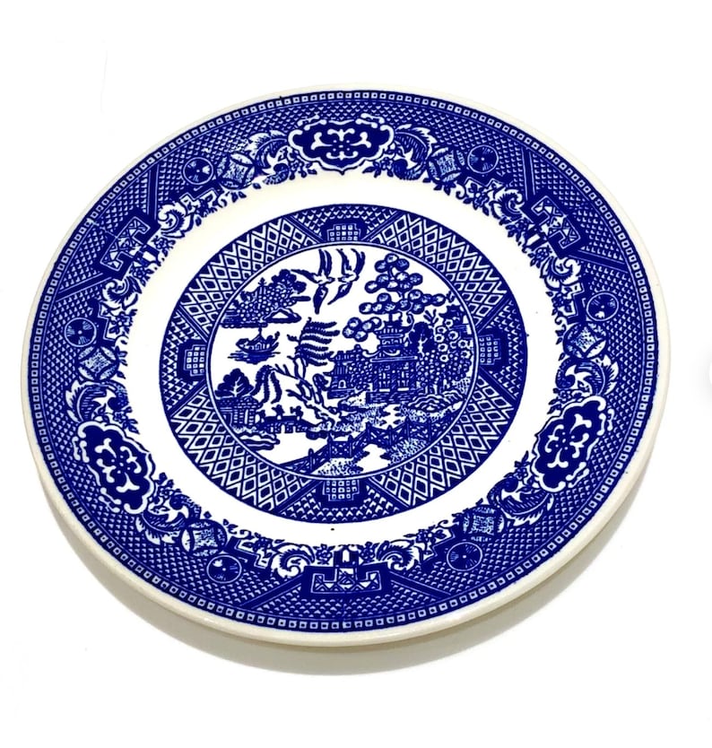 May include: A round, white and blue ceramic plate with a detailed Willow pattern. The plate features a central scene of a bridge, pagoda, and birds, surrounded by a geometric and floral border. The plate is approximately 25 cm in diameter.