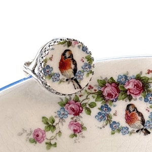 May include: A silver ring with a round, white face featuring a detailed painting of a bird with red, brown, and black feathers. The ring is set against a floral background with pink roses and blue flowers.