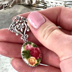K&ouml;nnte beinhalten: Silberfarbener Anh&auml;nger mit aufwendigem Filigran-Design. Er hat ein ovales Porzellan-Cameo mit einem floralen Rosendesign in Rot, Rosa und Gelb auf cremefarbenem Hintergrund. Der Anh&auml;nger wird in einer Hand gehalten.