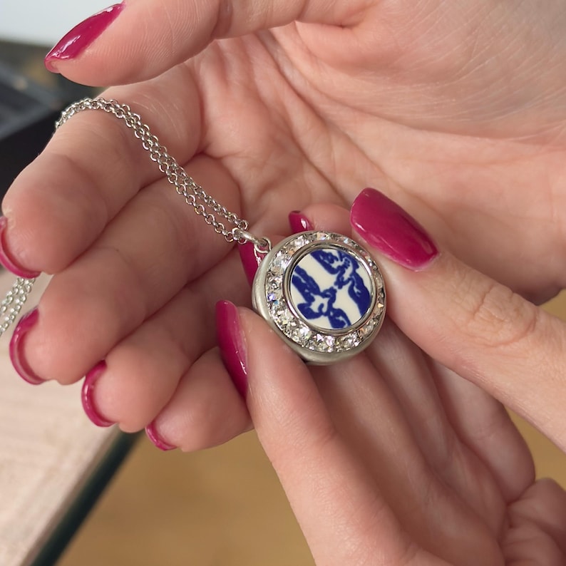 May include: A silver necklace with a circular pendant. The pendant has a blue and white pattern, encircled by clear crystals. The necklace is held by a person with red nail polish.