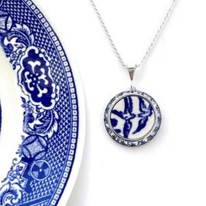 May include: A silver necklace with a circular pendant showcasing a blue and white porcelain pattern. The pendant is encircled by small, clear crystals. A section of a blue and white patterned plate is in the background.