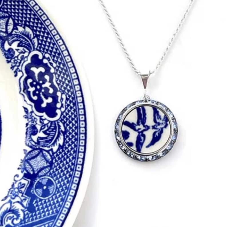 May include: A silver necklace with a circular pendant showcasing a blue and white porcelain pattern. The pendant is encircled by small, clear crystals. A section of a blue and white patterned plate is in the background.