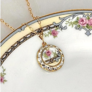 May include: A gold-toned necklace with a round pendant featuring a floral design with pink roses. The pendant is encircled by clear crystals. The necklace rests on a decorative plate with floral and geometric patterns.