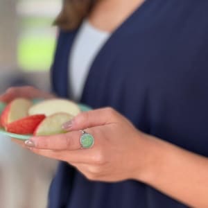 May include: A silver ring with a large, round, light green stone. The ring is on a person's finger, and the person is holding a plate of apple slices. The ring has a decorative border.