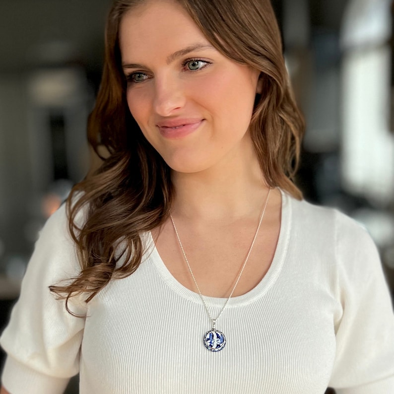 May include: A silver necklace with a circular pendant displaying a blue and white pattern. The pendant is suspended from a fine chain. The woman is wearing a white top.