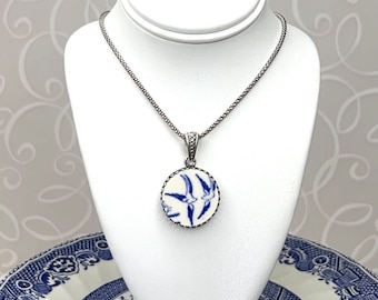 May include: A round pendant necklace featuring a white ceramic piece with a blue bird pattern, set in a silver-toned metal frame. The pendant hangs from a textured silver-toned chain, showcasing a detailed design of flying birds in blue.