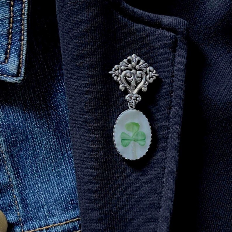 K&ouml;nnte beinhalten: Silberfarbene Brosche mit einem verzierten Oberteil und einem ovalen Anh&auml;nger. Der Anh&auml;nger zeigt ein gr&uuml;nes Kleeblatt-Design auf wei&szlig;em Hintergrund. Die Brosche ist an einer marineblauen Jacke befestigt.