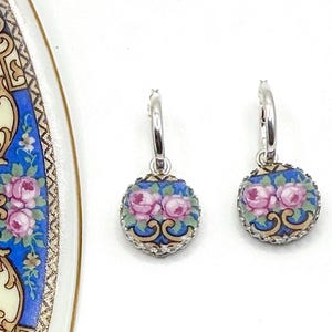 Peut inclure: Une paire de boucles d'oreilles cr&eacute;oles en argent avec des pendentifs circulaires sur fond bleu, orn&eacute;s de motifs de roses roses et de volutes dor&eacute;es. Les boucles d'oreilles sont pr&eacute;sent&eacute;es &agrave; c&ocirc;t&eacute; d'un fragment d'assiette en c&eacute;ramique.