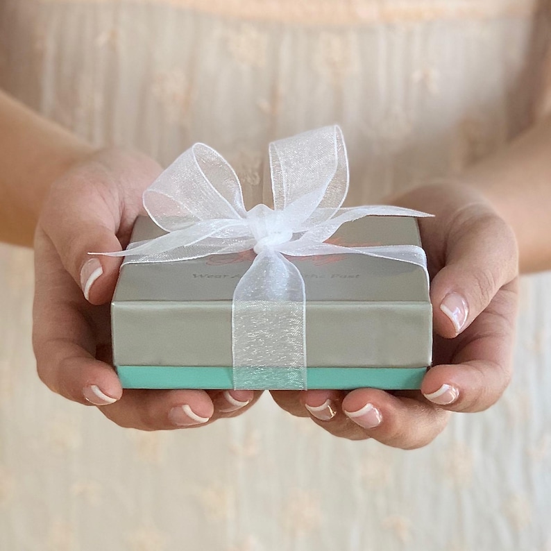 May include: A small, rectangular gift box with a light grey top and a turquoise base, tied with a sheer white ribbon and bow. The box is held in open hands against a soft, blurred background.