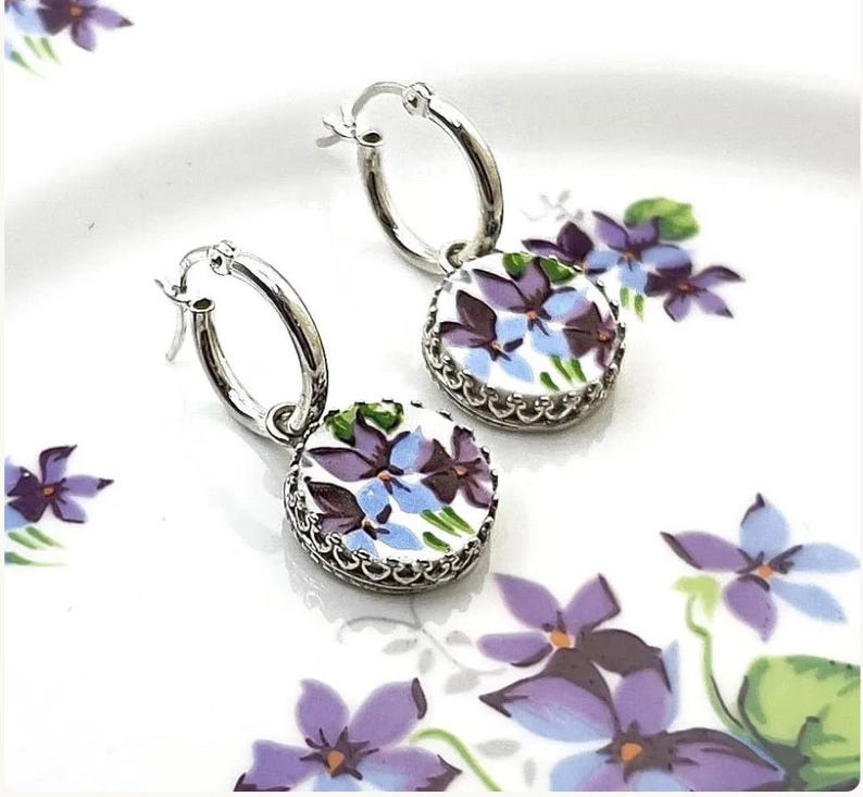 Peut inclure: Une paire de boucles d'oreilles cr&eacute;oles en argent avec des breloques rondes blanches orn&eacute;es d'un motif floral de violettes violettes et bleues. Chaque breloque est ench&acirc;ss&eacute;e dans un serti en argent festonn&eacute;, suspendue &agrave; un anneau.