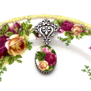 K&ouml;nnte beinhalten: Ein silberfarbener Anh&auml;nger mit ovalem Blumendesign. Der Anh&auml;nger zeigt rote, rosa und gelbe Rosen mit gr&uuml;nen Bl&auml;ttern. Er h&auml;ngt an einem dekorativen silbernen Filigranelement. Der Hintergrund ist ein wei&szlig;er Teller mit floralem Muster.