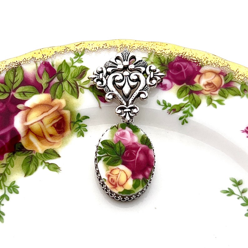 K&ouml;nnte beinhalten: Ein silberfarbener Anh&auml;nger mit ovalem Blumendesign. Der Anh&auml;nger zeigt rote, rosa und gelbe Rosen mit gr&uuml;nen Bl&auml;ttern. Er h&auml;ngt an einem dekorativen silbernen Filigranelement. Der Hintergrund ist ein wei&szlig;er Teller mit floralem Muster.