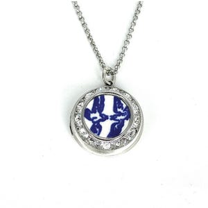May include: A silver locket necklace with a circular pendant. The pendant displays a blue and white floral pattern, encircled by clear crystals. The necklace is suspended from a fine chain.