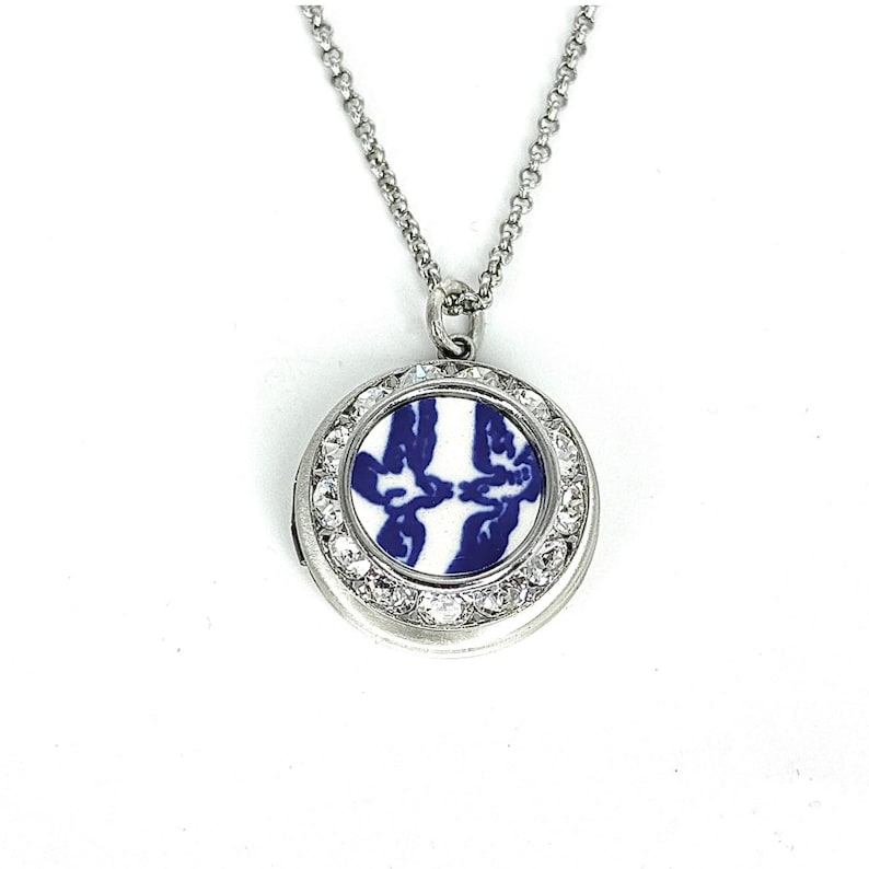 May include: A silver locket necklace with a circular pendant. The pendant displays a blue and white floral pattern, encircled by clear crystals. The necklace is suspended from a fine chain.